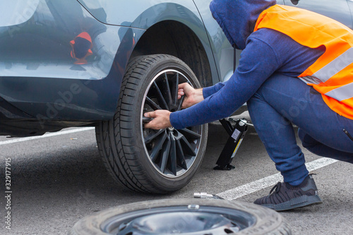 A man in a orange safety vest changes a flat tire on a road. Close-up mans hands to the wheel of a broken car. Replacement of a wheel using skrewdriver.