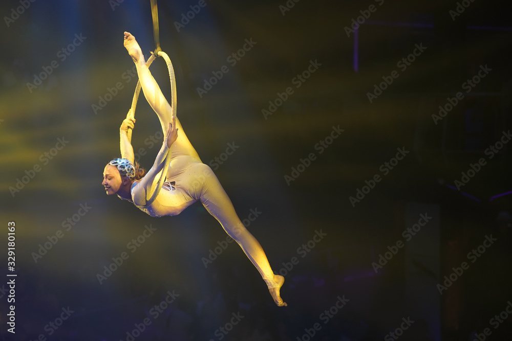 Obraz premium Flexible young woman make performance on aerial hoop, flexible back and split on aerial hoop, aerial circus show, yellow light. Flexible woman gymnast upside down on hoop. Show in night club. 