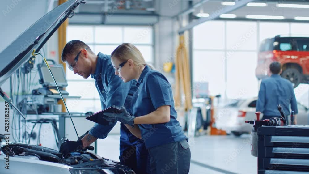 Two Mechanics in a Service are Inspecting a Car After They Got the Diagnostics Results. Female Specialist is Comparing the Data on a Tablet Computer. Repairman is Using a Ratchet to Repair the Faults.