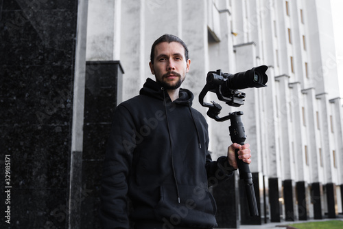 Young Professional videographer holding professional camera on 3-axis gimbal stabilizer. Pro equipment helps to make high quality video without shaking. Cameraman wearing black hoodie making a videos.