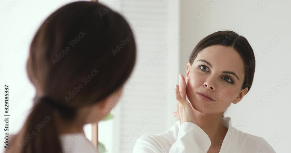 Confident beautiful young woman putting facial cream on looking in mirror. 20s girl applying face creme on flawless perfect healthy fresh skin. Skincare everyday treatment morning beauty concept Confident beautiful young woman putting facial cream on looking in mirror. 20s girl applying face creme on flawless perfect healthy fresh skin. Skincare everyday treatment morning beauty concept