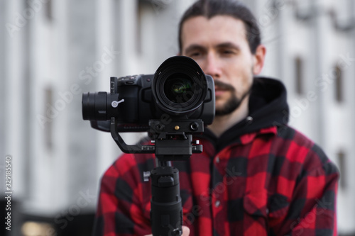 Young Professional videographer holding professional camera on 3-axis gimbal stabilizer. Pro equipment helps to make high quality video without shaking. Cameraman wearing red shirt making a videos. © Volodymyr
