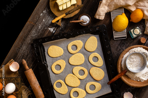 overhead shot of baked egg shaped easter cookies on baking tray on rustic wooden table with flour, eggs, butter, lemon. process of baking cookies. easter card