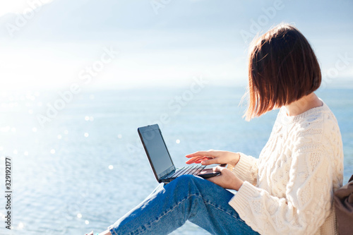 Young woman working at sea beach. Girl freelancer using laptop and the Internet in outdoor office in traveling. Workplace on pier. Successful female business, self development. Copy space.