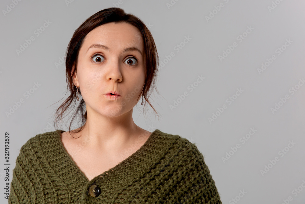 Close up view of surprised young brunette woman covering her wide-open mouth in shock and astonishment posing isolated against gray wall background. Human face expressions and emotions Close up view of surprised young brunette woman covering her wide-open mouth in shock and astonishment posing isolated against gray wall background. Human face expressions and emotions