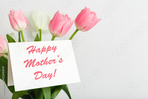 Happy Mothers Day text on gift card in flower bouquet on white background. Greeting card for Mom. Flower delivery, Congratulations card in flowers for women. Greeting card in pink tulips.
