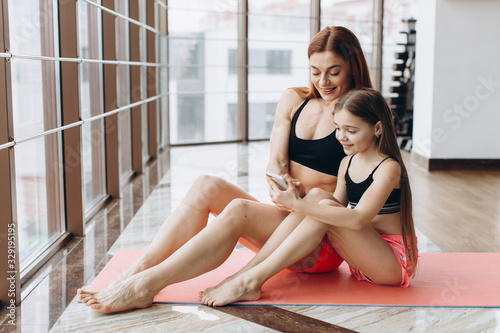 Mother and daughter relaxing after workout sitting on mat, using phone in gym for watching video how do exercise