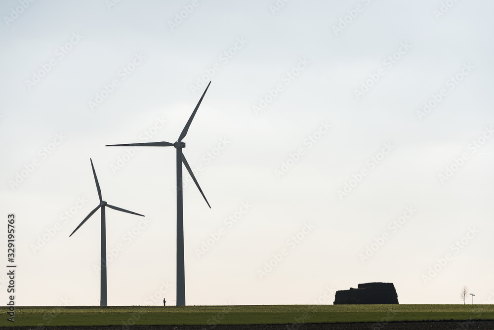 Fototapeta premium Two wind farms and a human silhouette showing the scale of these objects. All on the background of blue sky and green field.