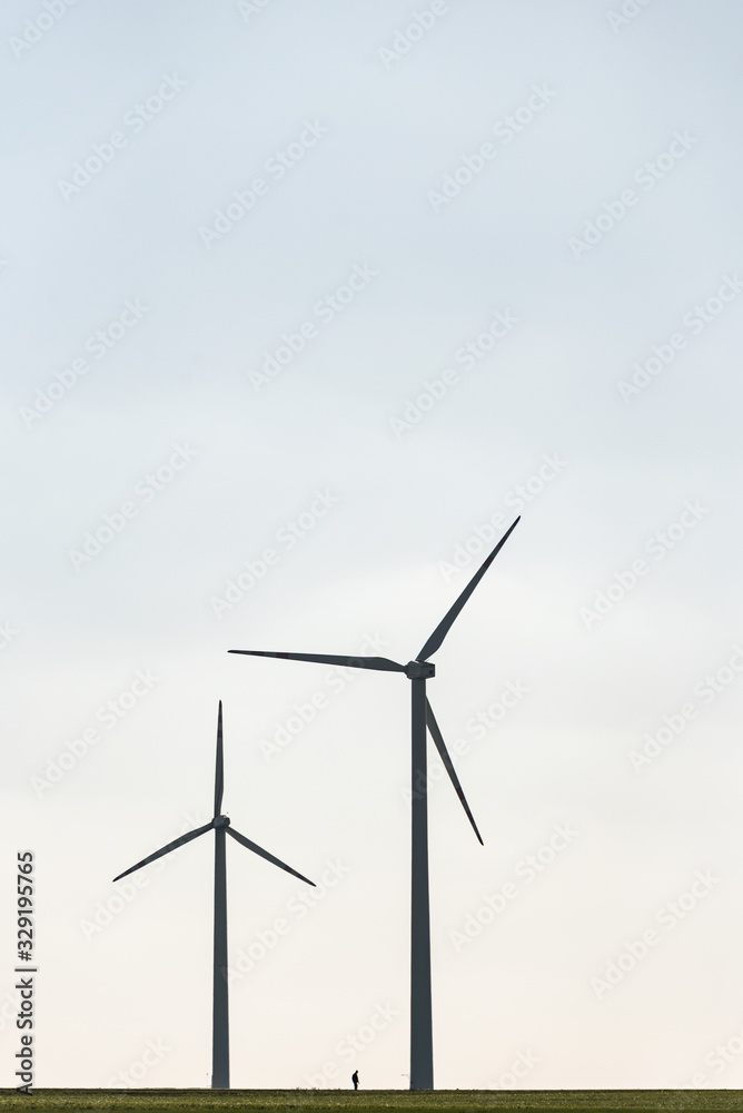 Two wind farms and a human silhouette showing the scale of these objects. All on the background of blue sky and green field. Two wind farms and a human silhouette showing the scale of these objects. All on the background of blue sky and green field.