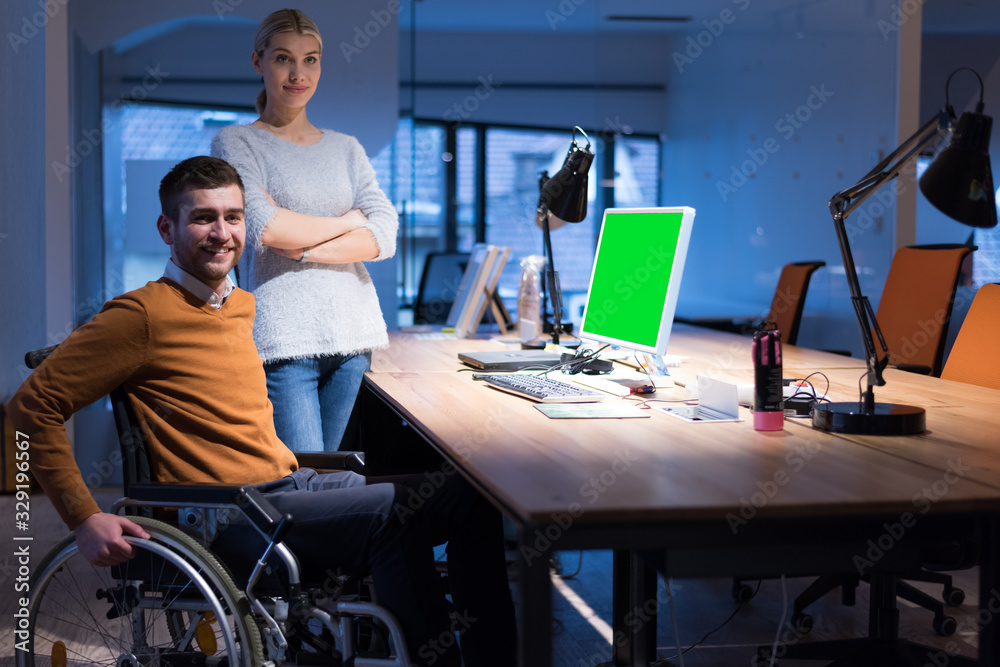 Handicapped young man with female colleague working in office. Disabled businessman in the wheelchair works in the office at the computer while performing in co-working space. Looking at camera. Handicapped young man with female colleague working in office. Disabled businessman in the wheelchair works in the office at the computer while performing in co-working space. Looking at camera.