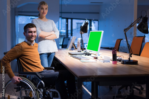 Handicapped young man with female colleague working in office. Disabled businessman in the wheelchair works in the office at the computer while performing in co-working space. Looking at camera.