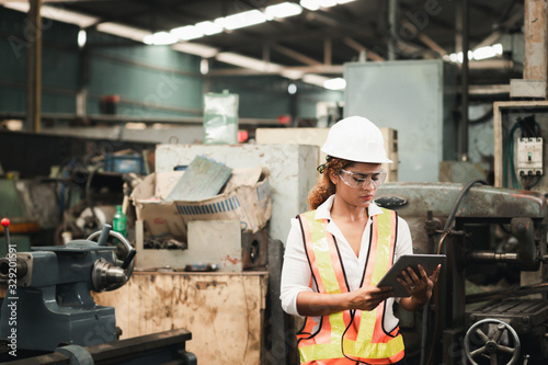 Female industrial engineer wearing a white helmet while standing in a heavy industrial factory behind she looking of working at industrial machinery and check security system setup in factory.