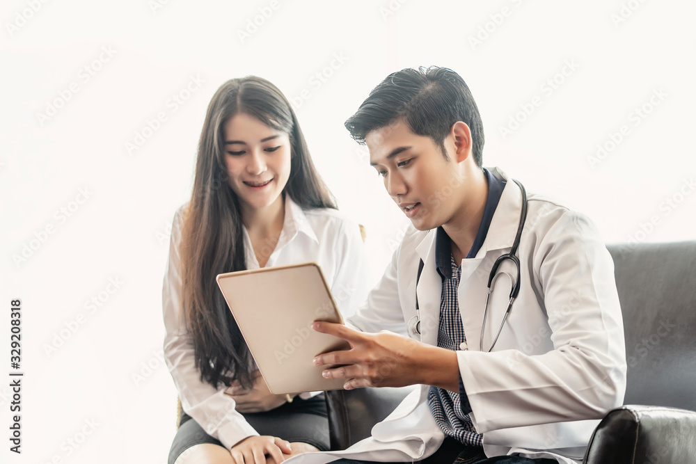 asian male doctor wearing a white scrub lab coat with a stethoscope around his neck smiling and holding a tablet showing health information to a young beautiful asian female patient smartly dressed. asian male doctor wearing a white scrub lab coat with a stethoscope around his neck smiling and holding a tablet showing health information to a young beautiful asian female patient smartly dressed.