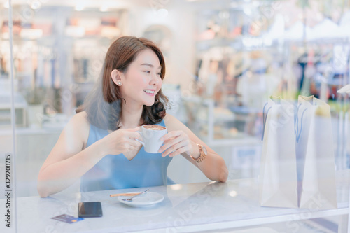 Free Download Asian woman enjoying coffee in a coffee shop viewed through the glass with reflections she sits at a table chatting and laughing happy after shopping with a shopping bag  in the cafe - GFXLoad