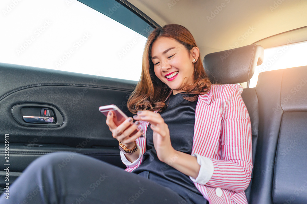 asian businesswoman smiling joyfully holding and playing on her smartphone while sitting in the backseat of a car, wearing a pink and white blazer, with sunset shining through the window background