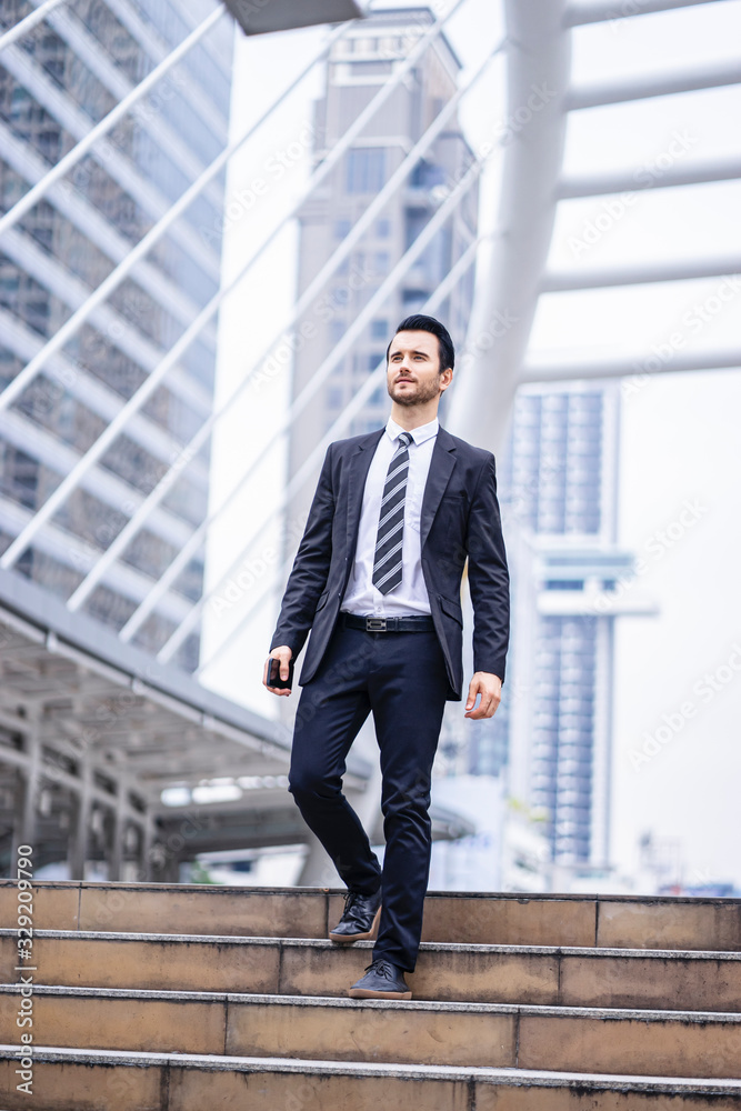 Caucasian handsome businessman confidently walking down the stairways while holding a smartphone, wearing black suit and tie with city skylines and urban architectural structure in the background