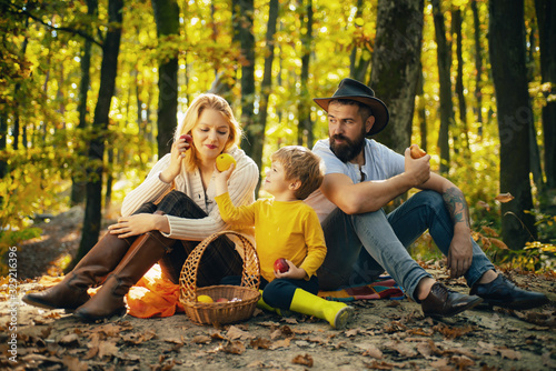 Happy family of three lying in the grass in autumn. A young family with small child having picnic in autumn nature at sunset. Mother, bearded father and kid are eating apples.