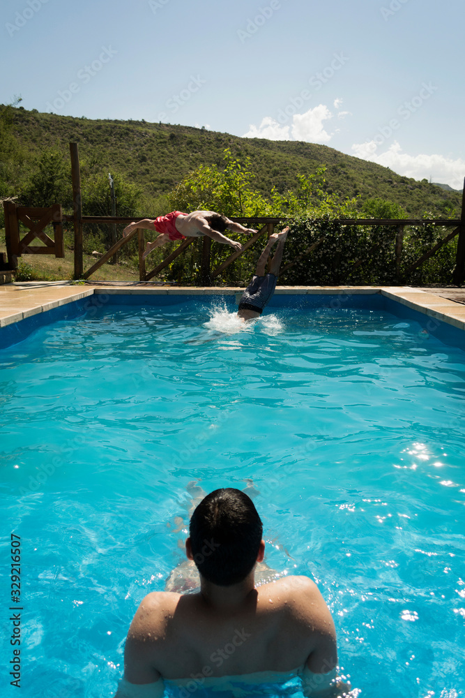 Two boys jumping into the pool while another boy on his back watches them