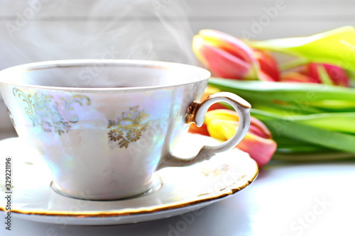 White porcelain pattern cup of hot black tea on dish with smoke on white desk with blurred Bouquet of flowers of red yellow blooming tulips with green leaf on backdrop. Selective focus