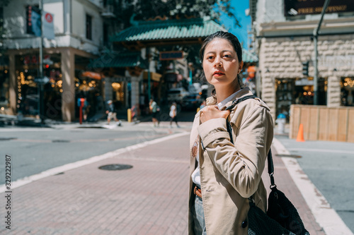 Young beautiful cute girl in stylish coat with fashionable bag on sunshine. charming lady local lifestyle concept. pensive pretty woman standing on sunny day street and looking front thoughtful.