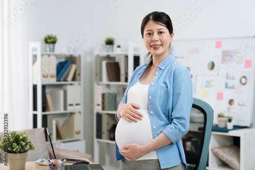 Cheerful pregnant lady freelancer working in modern home office in day time. beautiful future motherhood standing indoors in bright workplace face camera smiling. maternity woman hold big belly.