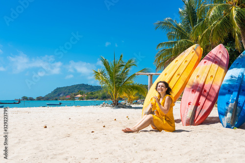Girl in a yellow dress on a tropical sandy beach works on a laptop near kayaks and drinks fresh mango. Remote work, successful freelance. Works on vacation.