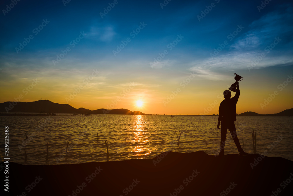 The shadow of the silhouetted man is happy and holds the trophy in the morning sun.