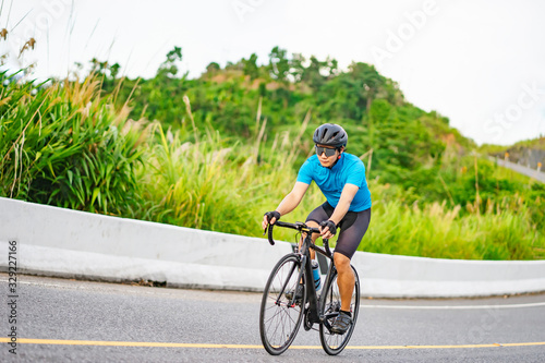 asian male riding on a black bicycle along the road, smiling and wearing a cycling blue jersey, crash helmet and goggles, on a long winding road with forest trees and mountains in the background.