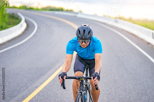 Fototapeta Naklejka Na Ścianę i Meble -  asian male riding on a black bicycle along the road, smiling and wearing cycling blue jersey, crash helmet and goggles, on a long winding road with forest trees and sun shining through the background.