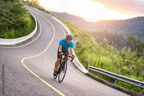 asian male riding on a black bicycle along the winding road up a hill, wearing a cycling blue jersey, crash helmet and goggles, sunset light, grey sky, and forest trees and mountains in the background