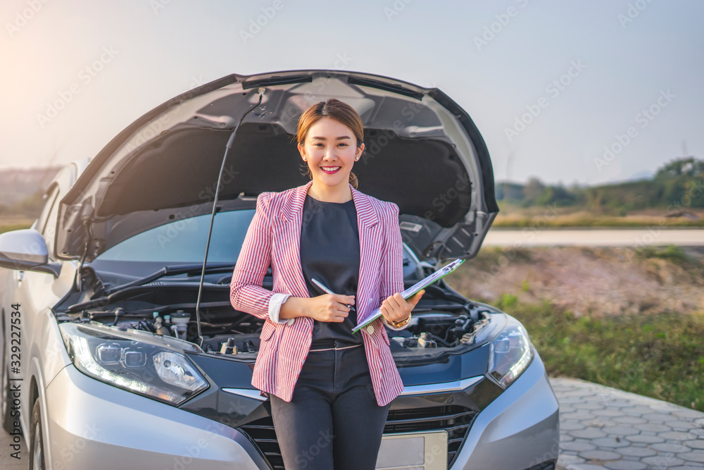 Naklejka premium portrait of asian female car inspector holding a pen clipboard checking on the engine with the paper work on the clipboard with the car hood opened up revealing the car engine, leaning against the car