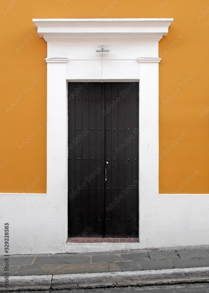 Fototapeta premium Weathered wooden doors set into an orange wall with white wooden trim along cobblestone sidewalk in old town San Juan.