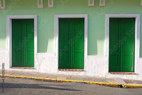 Three green doors trimmed in white set into a mint green paint wall on a grey brick sidewalk