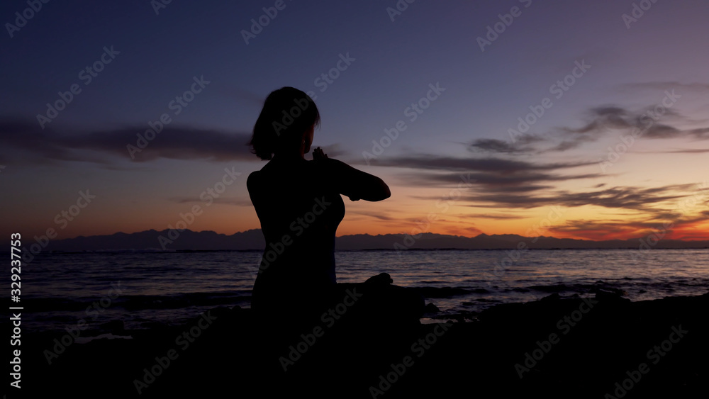 Silhouette of girl sits in namaste pose meditating by the sea at sunset.