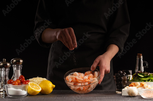 Chef cooks shrimps, sprinkled with salt, freezing in motion. Against the background of vegetables, Seafood, healthy seafood. Black background for design