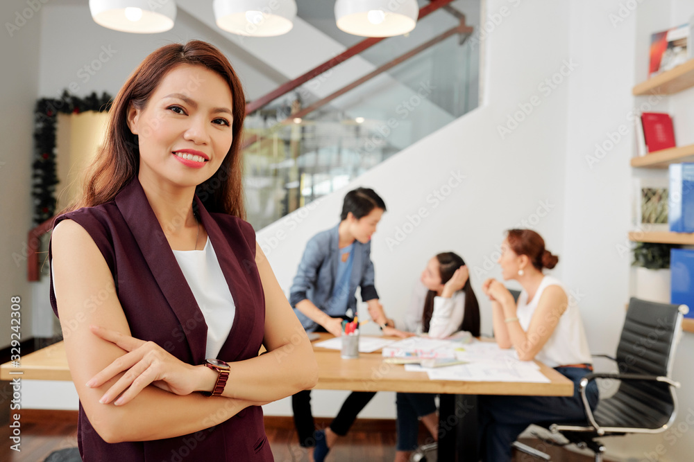 Pretty young confident businesswoman folding arms and smiling at camera when her team discussing project ideas Pretty young confident businesswoman folding arms and smiling at camera when her team discussing project ideas