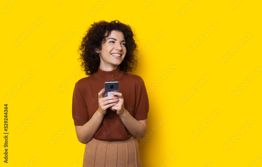 Stunning curly haired caucasian girl is smiling on a yellow wall while texting with somebody