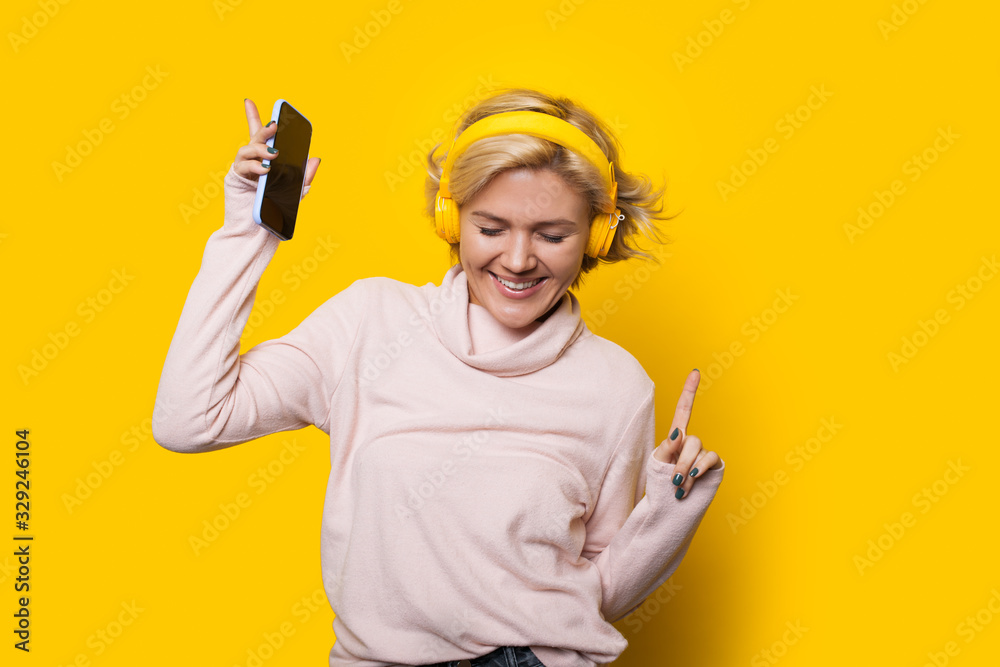 Smiling caucasian girl with blonde hair is dancing on a yellow background while listening to music through headphones Smiling caucasian girl with blonde hair is dancing on a yellow background while listening to music through headphones