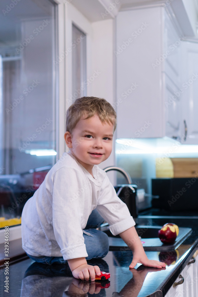 Obraz premium Portrait of happy joyful beautiful boy in kitchen background. Child eats an apple, rolls a toy car on the table and enjoys life. happy and carefree childhood. Healthy eating concept.