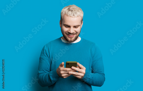 Caucasian man with blond hair and beard is wearing a sweater and typing a message on phone posing on a blue background © Strelciuc