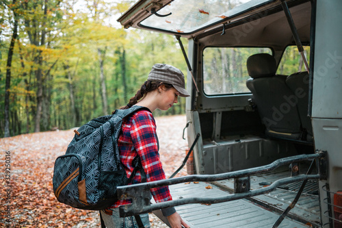 A young Caucasian woman with a backpack on her back closes the trunk of a jeep. In the background, trees and forest. Close up. Concept of car travel and tourism
