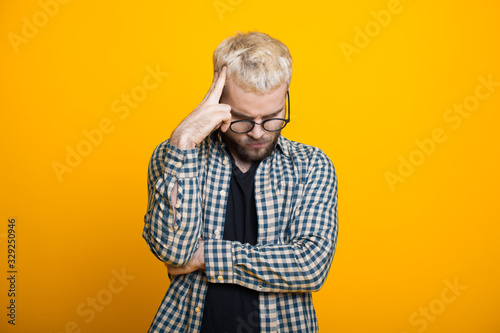 Caucasian man with blonde hair and beard is touching his head while thinking about something and posing on a yellow background