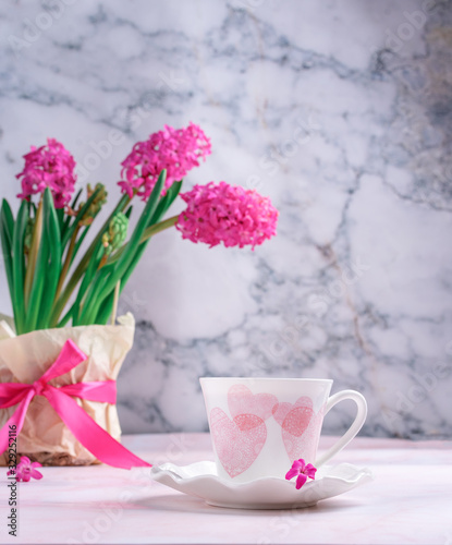 white cup with pink hearts with a vase of spring flowers on the table against the background of gray marble. Morning coffee, girl’s breakfast, spring mood