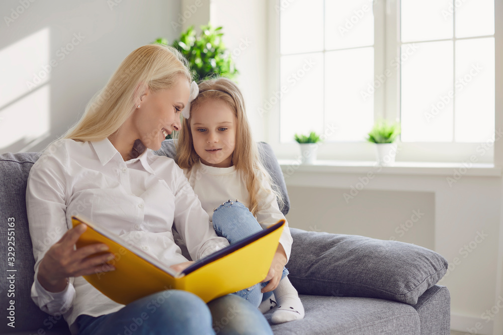 Mother and daughter are reading a book while sitting on a sofa in a room with a window. Mother and daughter are reading a book while sitting on a sofa in a room with a window.