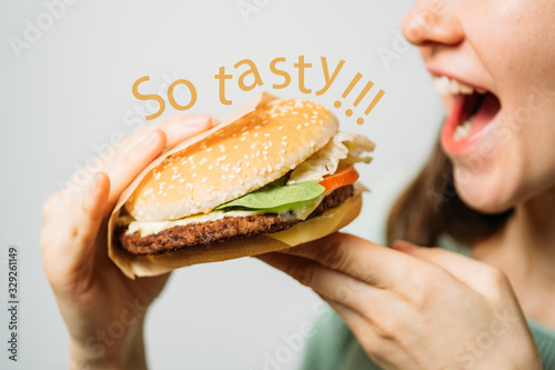 The girl holds a hamburger with her hands and eats it with appetite. The inscription is so tasty.