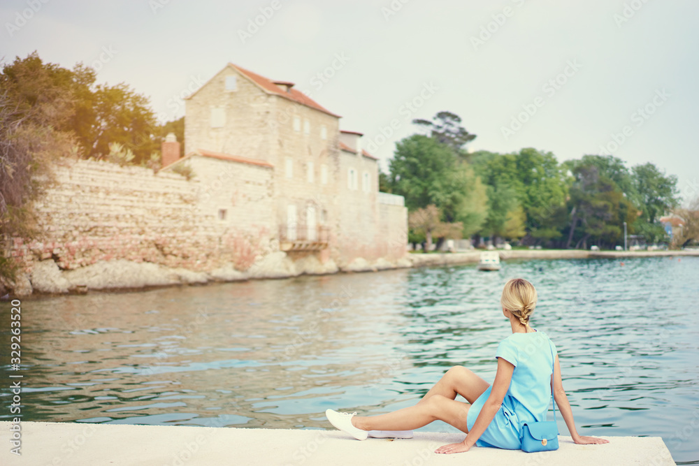 Tourism concept. Young traveling woman enjoying the view of Kastel Castle sitting near the sea on Croatian coast.