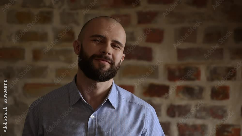 A close portrait of a serious office worker who starts smiling and looks straight at the camera. A man with a beard wears two silver earrings and a blue shirt. A close portrait of a serious office worker who starts smiling and looks straight at the camera. A man with a beard wears two silver earrings and a blue shirt.