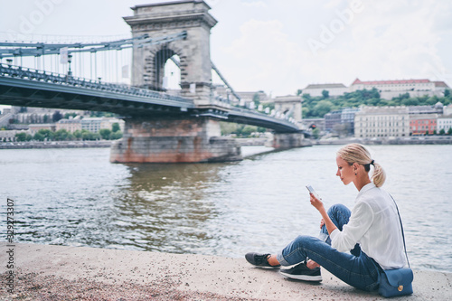 Vacation in Budapest. Young traveling woman using smartphone on riverside promenade with city view. © luengo_ua