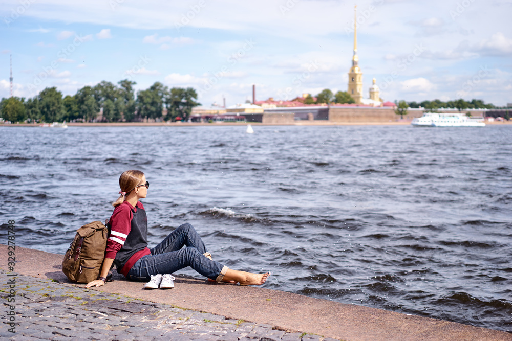 Travel and tourism. Young woman sitting on embankment of Neva river enjoying the view of Peter and Paul fortress, Saint Petersburg, Russia.