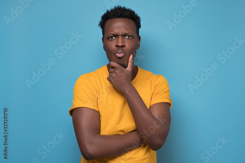 Thoughtful African american man in yellow clothes and glasses thinking or planing how to become a businessman. Studio shot on blue wall.